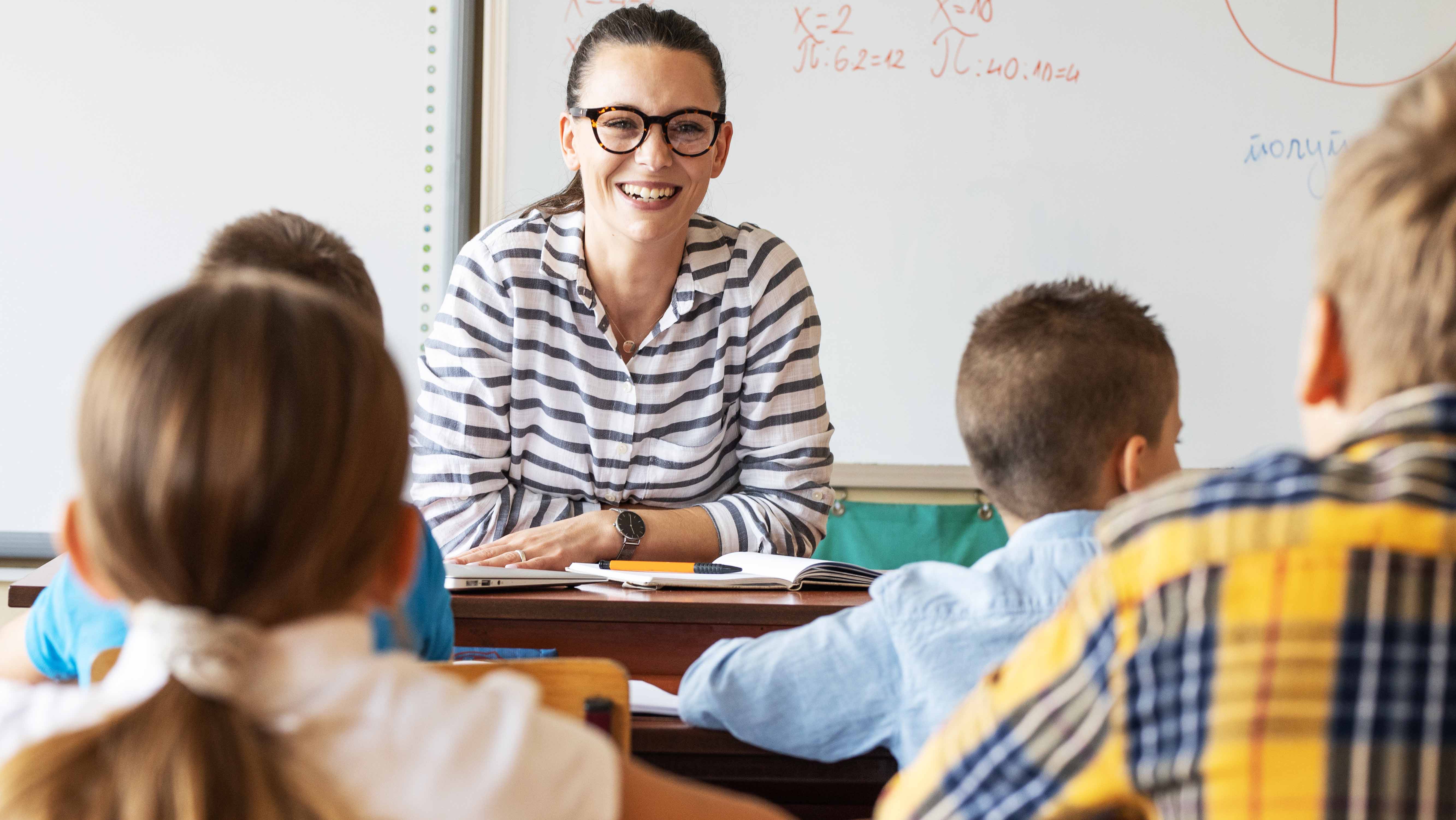 Teacher seated at desk in front of class smiling while lecturing