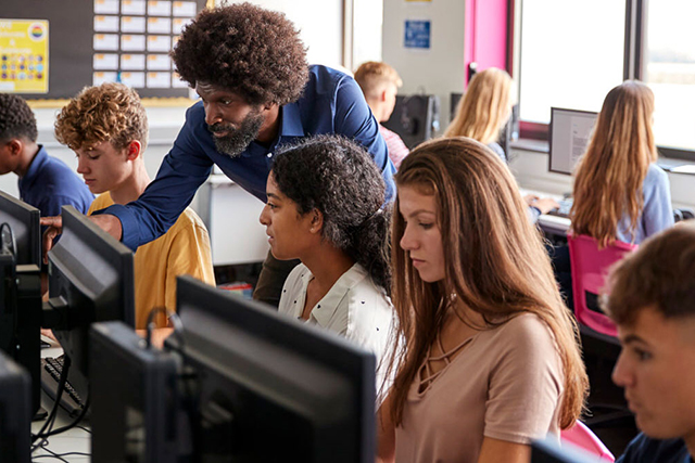 A teacher helps high school students sitting at computers in a classroom. 