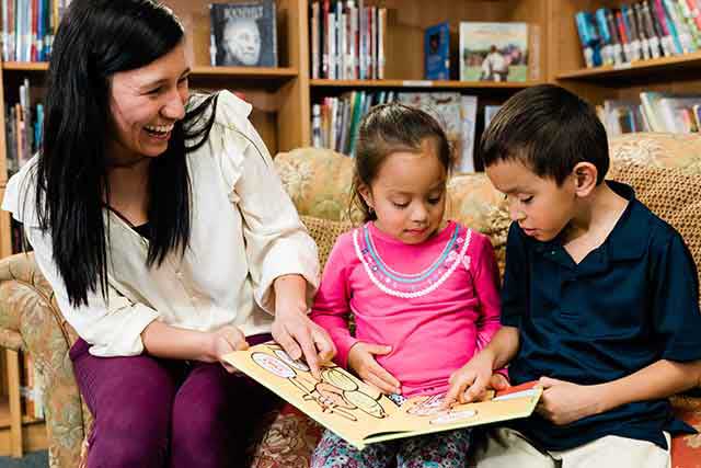 A teacher points to pictures in a children's book as she smiles at two young children sitting on a couch in a library at a public school.