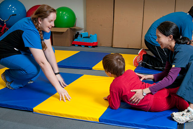 A young boy lies face down on colorful mats and is helped by an adult to crawl to another woman.