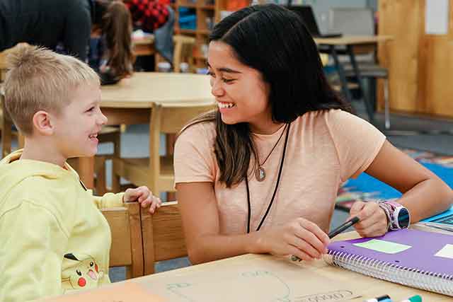 A UNT student teacher smiles at a young boy as they sit together at an art table. 