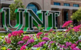 Late summer flowers bloom outside of the UNT Welcome Center with the UNT sculpture in the background.